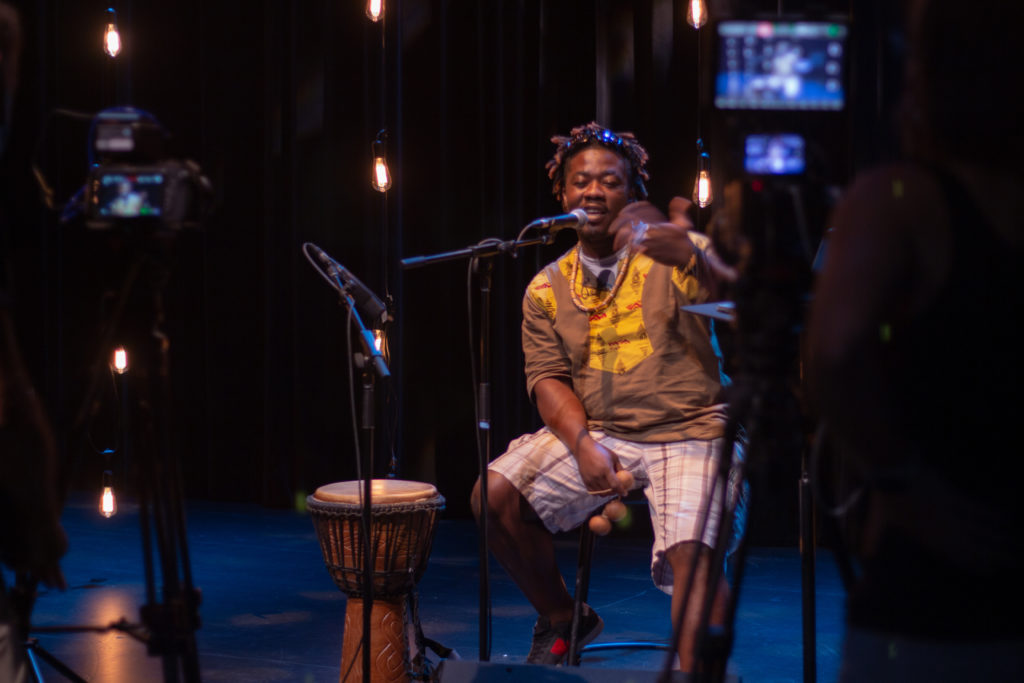 A seated Black man is filmed as he speaks into a microphone, smiling, holding percussion balls, a hand drum next to him. / Un homme Noir assis est filmé tans qu’il parle dans un microphone en souriant et en tenant des boules percutantes, un tambour à main à ses côtés.