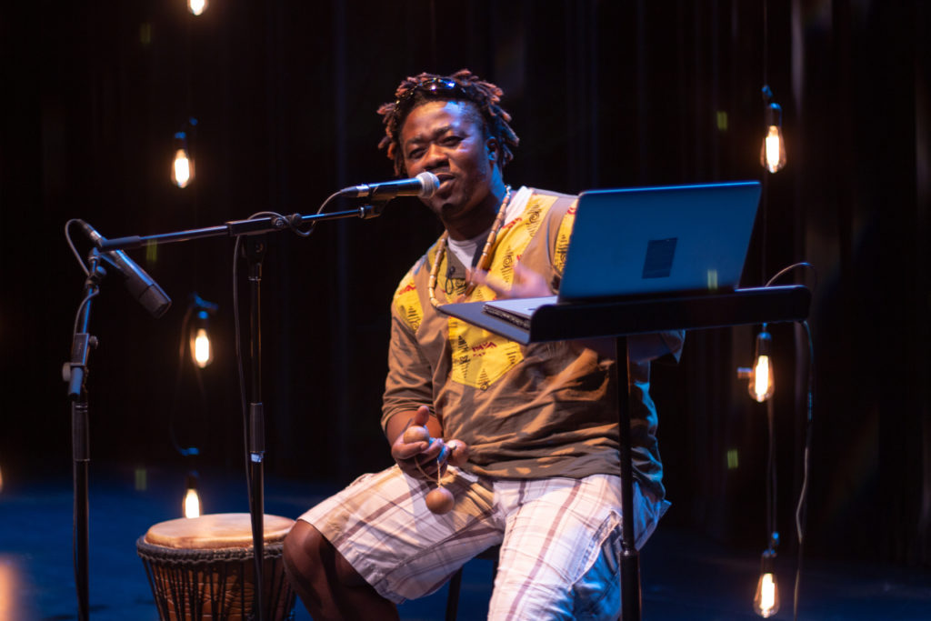 A Black man speaks into a microphone, smiling, holding percussion balls, a hand drum on one side of him, an open laptop on the other. / Un homme Noir parle dans un microphone en souriant et en tenant des boules percutantes, un tambour à main d’un côté, un ordinateur portable ouvert de l’autre.