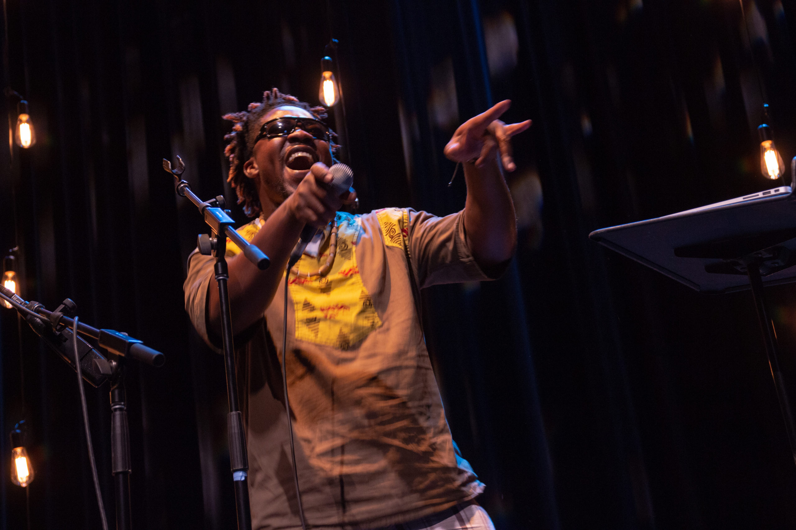 A smiling Black man with sunglasses on holds out a microphone to the audience as he points at them. / Un homme Noir souriant en lunettes de soleil tient son microphone vers ses spectateurs et les pointe du doigt.