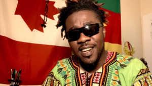 A Black man wears sunglasses and a multicoloured patterned shirt in front of Canadian and Cameroonian flags. / Un homme Noir porte des lunettes de soleil et un chandail multicolores à motifs devant des drapeaux canadiens et camerounais.