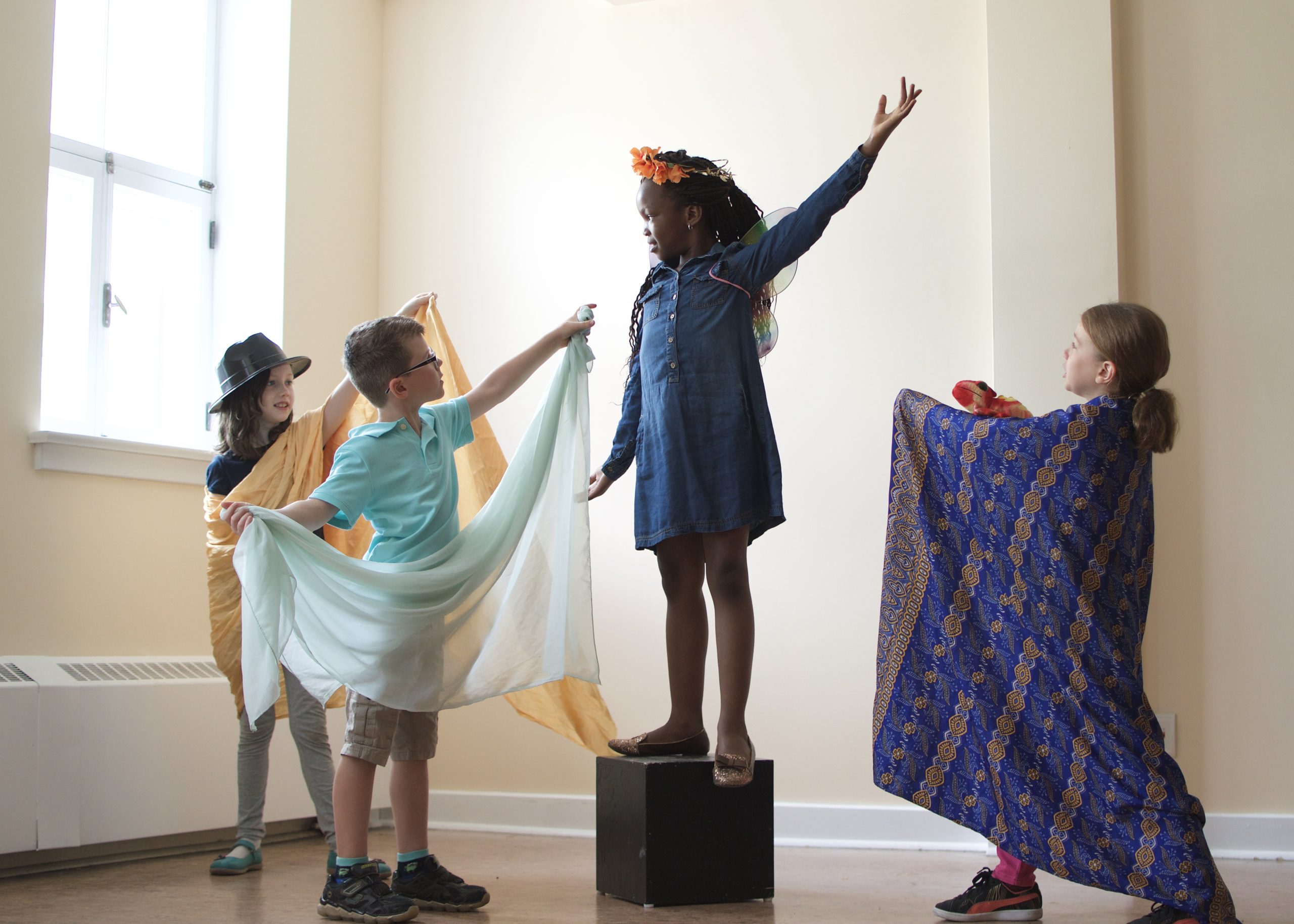 Four children in costumes act; three of them holding their arms out to the fourth who stands on a box, one arm towards the sky. / Quatre enfants en costumes jouent; trois d’entre eux tiennent leur bras dans la direction de la quatrième qui est debout sur un cube, un bras vers le ciel.