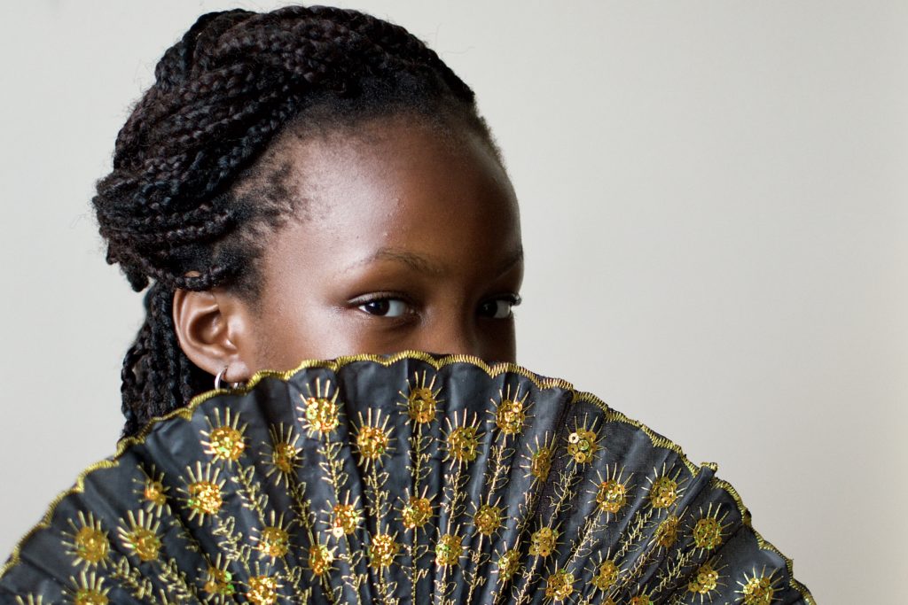 A little Black girl hides half her face with a gold sequinned black fan. / Une petite fille Noire se cache la moitié du visage avec un éventail noir aux paillettes dorées.