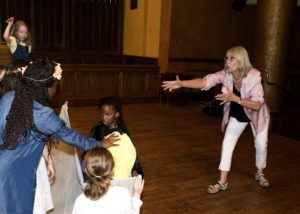 A White woman in a theatre passionately explains something to a group of children in front of her. / Une femme Blanche dans un théâtre explique quelque chose passionnément à un groupe d’enfants devant elle.