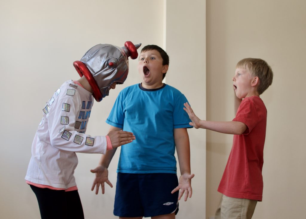 A child wearing a robot costume acts like one while two boys watch, scared. / Un enfant porte un costumer de robot et en imite un tandis que deux garçons le regardent, apeurés.