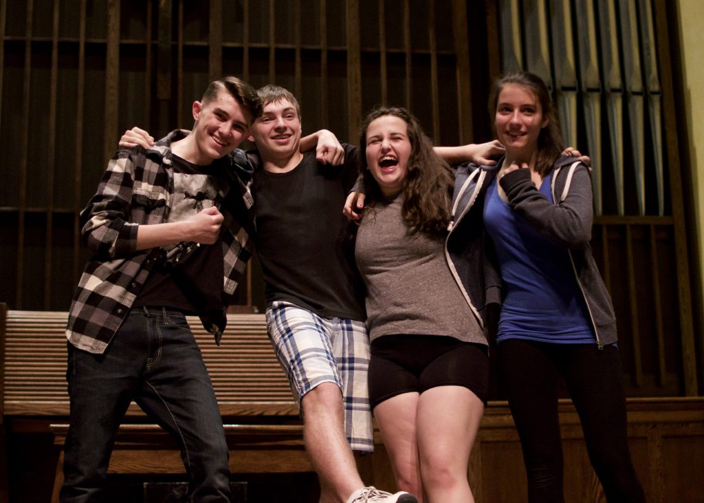 Four White teenagers pose with their arms around each other while laughing. / Quatre adolescents Blancs posent, les bras autours des autres, en rigolant.