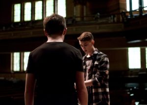 Two White people discuss on a stage in a theatre. / Deux personnes Blanches se parlent sur une scène dans un théâtre.