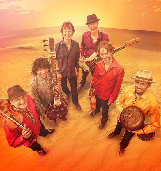 Six men smile, five of them holding their string instruments and one holding a drum, with a desert as the backdrop. / Six hommes sourient, cinq d’entre eux tiennent leurs instruments à corde et l’un d’entre eux tient un tambour, ayant un désert comme toile de fond.