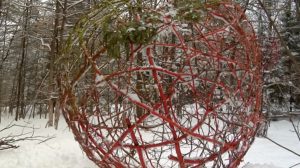 A large ball made of brown, green and red branches in a snowy forest. / Un grand ballon fait de bâtons marrons, verts et rouges dans une forêt enneigée.