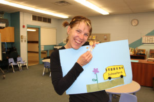 A smiling White woman in a classroom holds up a pale blue poster board with a flower, school bus and little girl glued on. / Une femme Blanche souriante dans une classe tient un poster bleu pâle avec collé dessus un autobus scolaire, une fleur et une petite fille.