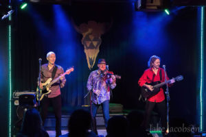 Three men play their string instruments on a stage: the electric guitar, the violin, and the flamenco guitar. / Trois hommes jouent leurs instruments à corde sur une scène: la guitare électrique, le violon et la guitare flamenca.