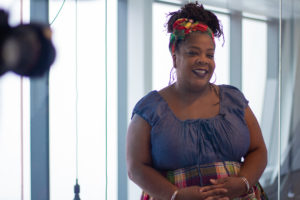 A Black woman smiles at the camera as another camera’s lens is pointed at her. / Une femme Noire sourit à l’appareil photo tandis que l’objectif d’une autre caméra se focalise aussi sur elle.