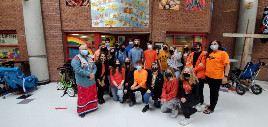A masked Indigenous Elder stands next to a group of masked teenagers and adults, most of whom wear orange shirts. / Une Aînée Autochtone portant un masque est à côté d’un groupe d’adolescents et d’adultes portant aussi des masques, la plupart d’entre eux portant aussi des hauts oranges.