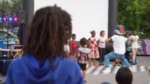 A young Black man in a blue hat dances outdoors in front of a small group of Black children as a person with dreads looks on. / Une personne avec des dreads observe un jeune homme Noir avec un chapeau bleu danser dehors devant un petit groupe enfants Noirs.