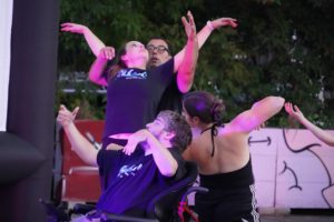 Two women and two men, one of whom is in a wheelchair, dance outside, basked in a pink light as a fifth person’s arm is in frame. / Deux femmes et deux hommes, don’t l’un en chaise roulante, dansent dehors sous une lumière rose alors que le bras d’une cinquième personne est aussi dans le cadre.