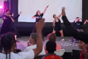 A man in a wheelchair and three women dance in front of a crowd, smiling, while another woman speaks into a microphone. / Un homme dans une chaise roulant et trois femmes dansent pour une foule en souriant pendant qu’une autre femme parle dans un microphone.