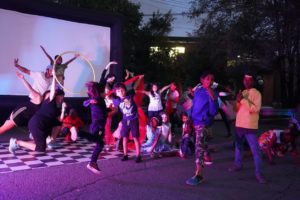 A group of children and a few adults pose outside at night, some holding red tulle or a hula hoop. / Un groupe d’enfants et quelques adultes pose dehors le soir, certains en tenant de la tulle rouge ou un cerceau.