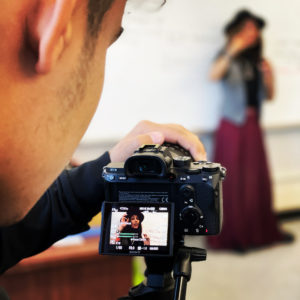 A young man in the foreground films a woman in front of a whiteboard on a digital camera, one hand on the lens. / Un jeune homme au le premier plan filme un femme devant un tableau sur une caméra digitale, une main sur l’objectif.