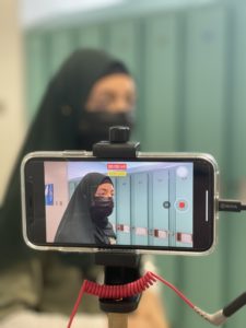 A young girl wears a Hijab and a mask in front of lockers as she is seen through the screen of an iPhone recording her. / Une jeune fille portant un Hijab et un masque devant des casiers est sur l’écran d’un iPhone qui la filme.