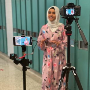 A young girl wearing a Hijab smiles as she is recorded on an iPhone and a Digital Camera, both on tripods. / Une jeune fille souriante portant un Hijab se fait filmer sur un iPhone et une Caméra digitale, les deux sur des tripodes.