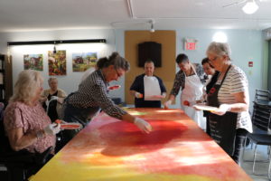 A group of seniors wearing aprons and white gloves gather around a long canvas that they paint red, yellow, and orange with sponges. / Un groupe de séniors avec des tabliers et des gants blancs sont autour d’un long tableau qu'ils peignent en jaune, rouge et orange avec des éponges.