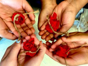 Four pairs of children’s hands each hold a small red fabric ball tied off with twine. / Quatre paires de mains d’enfants tiennent une petite boule de tissu rouge attaché avec de la corde.