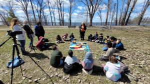 An Indigenous Elder stands outdoors in a circle of seated children, a blanket in the centre, as three adults look on. / Trois adultes écoutent une Aînée Autochtone dans un parc qui est debout, encerclée d’enfants assis, une couverture au centre.