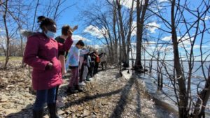 Children lined up next to each other hold one hand in the air as they stand on a rocky shore of the Ottawa River. / Une rangée d’enfants debout l’un à côté de l’autre sur une rive rocheuse de la Rivière des Outaouais lèvent une main en l’air.