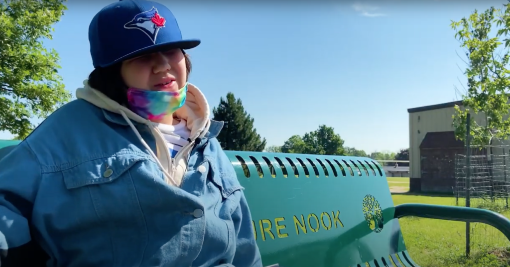 A young girl wearing a tie-dye mask just below her mouth and a BlueJays baseball hat that hides her eyes sits on a park bench. / Une jeune fille portant un masque tie-dye juste en dessous de sa bouche et une casquette des BlueJays qui cache ses yeux est assise sur un banc dans un parc.