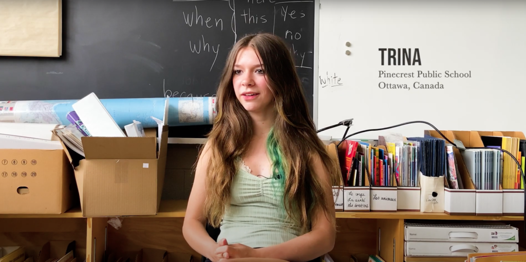 A young White girl with long brown and green hair sits in a classroom, in front of a whiteboard and a blackboard, the following text typed next to her head: TRINA, Pinecrest Public School, Ottawa, Canada. / Une jeune fille Blanche aux longs cheveux bruns et verts est assise dans une salle de classe devant deux tableaux, un texte tapé à côté de sa tête: TRINA, École publique Pinecrest, Ottawa, Canada.