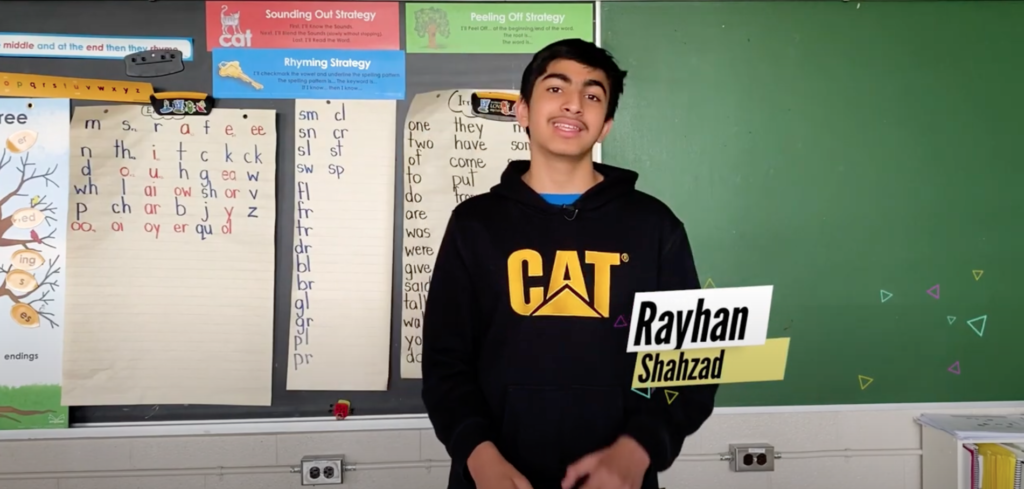 A young Brown boy with short black hair and a thin moustache stands in front of a blackboard in a classroom, a graphic in front of him states: Rayhan Shahzad. / Un jeune garçon basané aux courts cheveux noirs et une fine moustache est debout devant un tableau dans une salle de classe avec un texte sur l’image: Rayhan Shahzad.