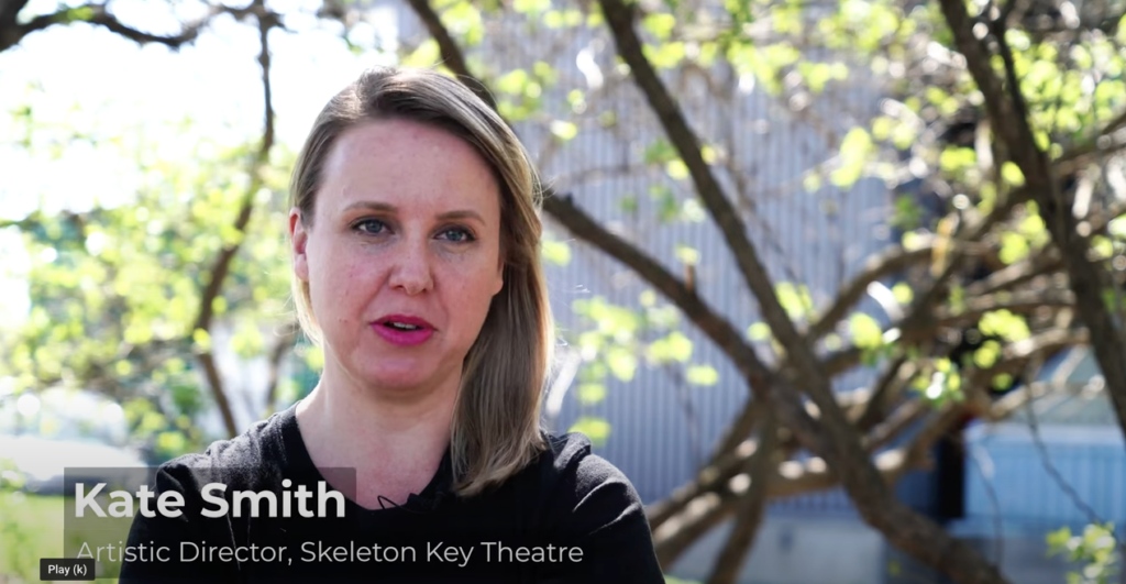 A White woman with blonde hair pushed to one side stands outside facing the camera with the follow text below her: Kate Smith, Artistic Director, Skeleton Key Theatre. / Une femme Blanche, ses cheveux blonds placés d’un côté, est dehors, le texte au bas de l’image indique: Kate Smith, Directrice Artistique, Skeleton Key Theatre.