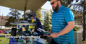 A White bearded man plays a drum kit made of recycled objects in a park with the following text at the bottom of the image: Junkyard Symphony, MASC Artist | Music, Parkdale Market Opening. / Un homme Blanc barbu joue de la batterie construite de bric-à-brac dans un parc avec du texte en bas de l’image qui se lit: Junkyard Symphony, Artiste MASC | Musique, Ouverture du Marché de Parkdale.