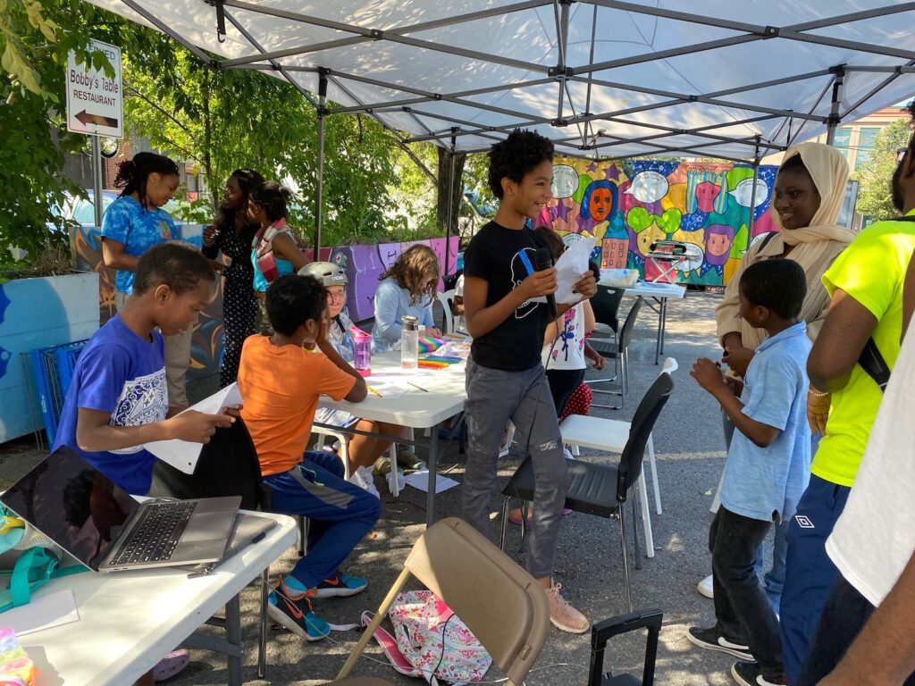 A group of children sit around a table outside drawing under a tent while others discuss around them. / Un groupe d’enfants est assis autour d’une table et dessine sous une tente alors que d’autres enfants discutent près d’eux.