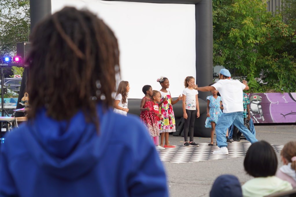 A young Black man in a blue hat dances outdoors in front of a small group of Black children as a person with dreads looks on. / Une personne avec des dreads observe un jeune homme Noir avec un chapeau bleu danser dehors devant un petit groupe enfants Noirs.