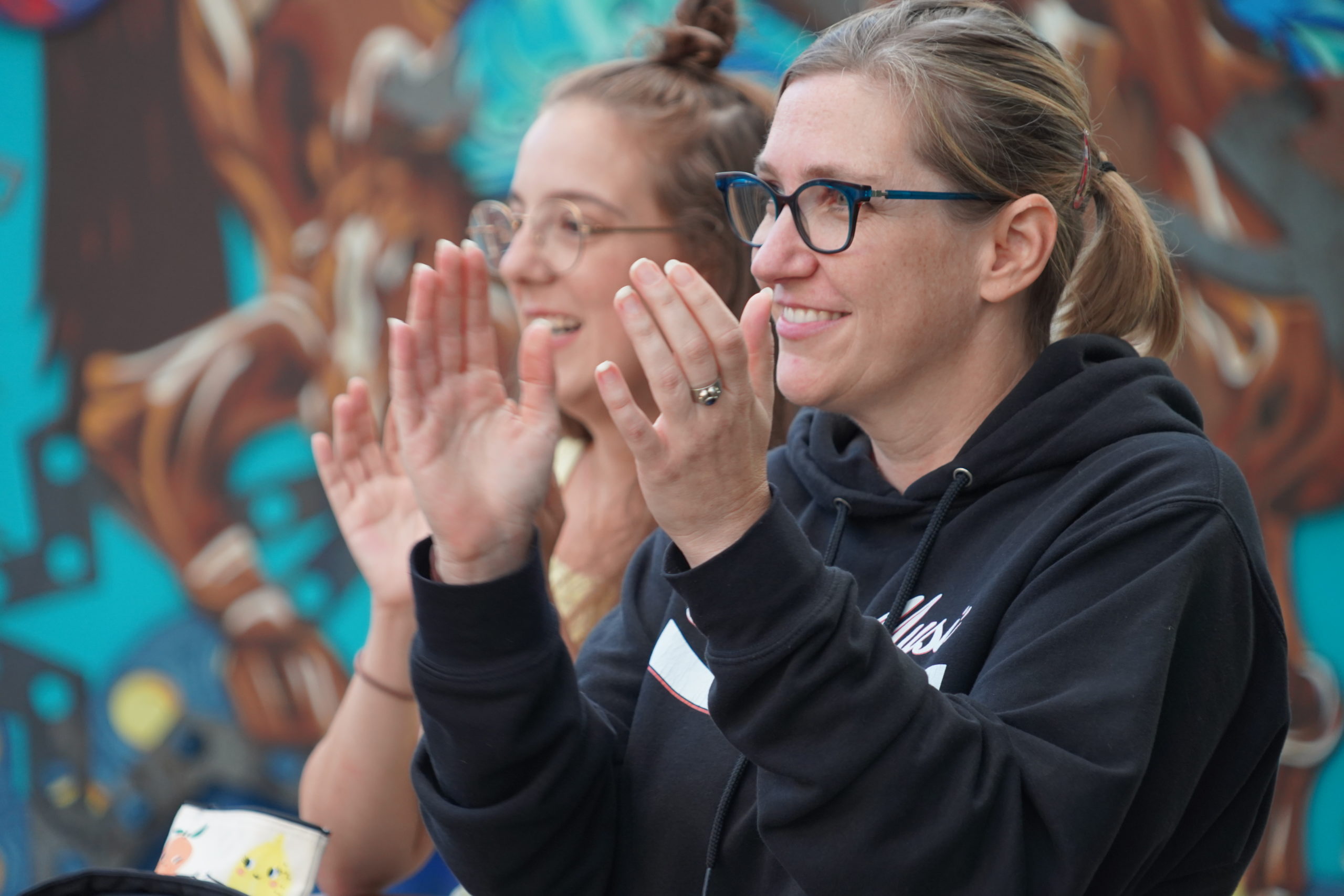 Two White women with glasses clap, their backs to a colourful mural. / Deux femmes Blanches avec des lunettes applaudissent, une murale colorée derrière elles.