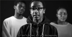 Black and white photo of young man in center, with a young man and woman in the background slightly blurred.