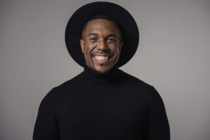 Stefan-Keyes, a CTV journalist, smiling against a grey backdrop. He is wearing a black turtle neck and a black wide rimmed hat.