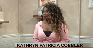 Photo of Arts educator for MASC name Kathryn Patricia Cobbler on Rogers TV. Patricia is wearing a pink blouse with a black undershirt and she has long brown curls.