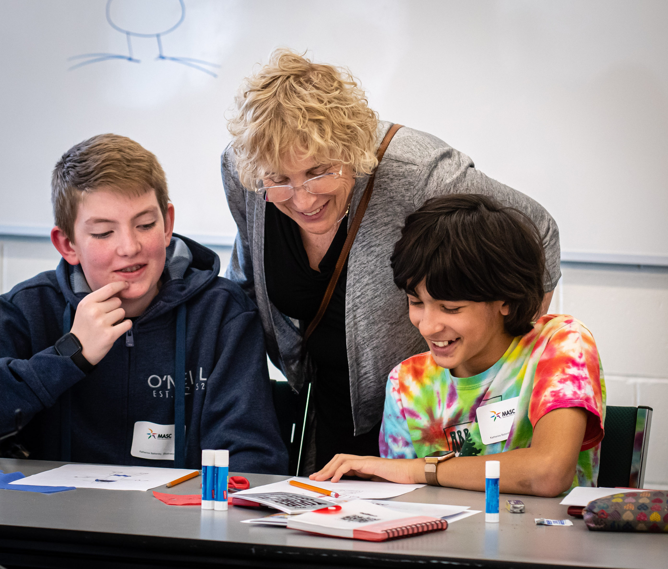 Roslyn Bern standing and leaning over a table with two boys on either side of her. The boys are smiling looking at a page.