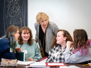 Roslyn Bern, president of the Leacross Foundation, standing and leaning forward at a table with 4 young women who are smiling. There are books and notebooks in front of them.