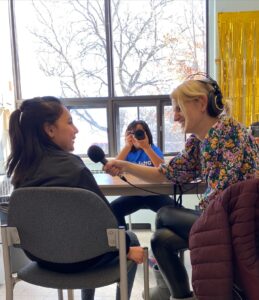 Olivia Robinson, a cbc reporter, is smiling holding a microphone in front of a girl with a brown hair in a half up pony tail. They are both sitting and there is another girl holding a camera behind them.