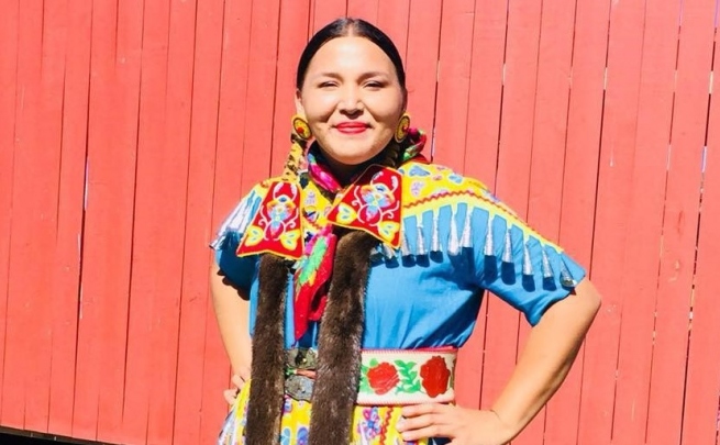 Amanda Fox looking forward proudly smilling outdoors on a sunny day. Her clothing is colourful in yellows, reds, and blues, and has traditional floral designs. There are tassels across her shoulders and her earrings are large yellow circles with a large red bead in the center. She looks radiant!