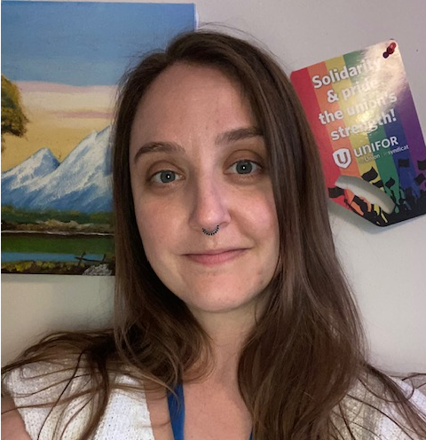 Megan Wegner, a community support outreach coordinator, smiling while looking forward. She has long light brown hair and a septum nose ring. There is a pride poster and a painted mountain landscape poster behind her on the wall.